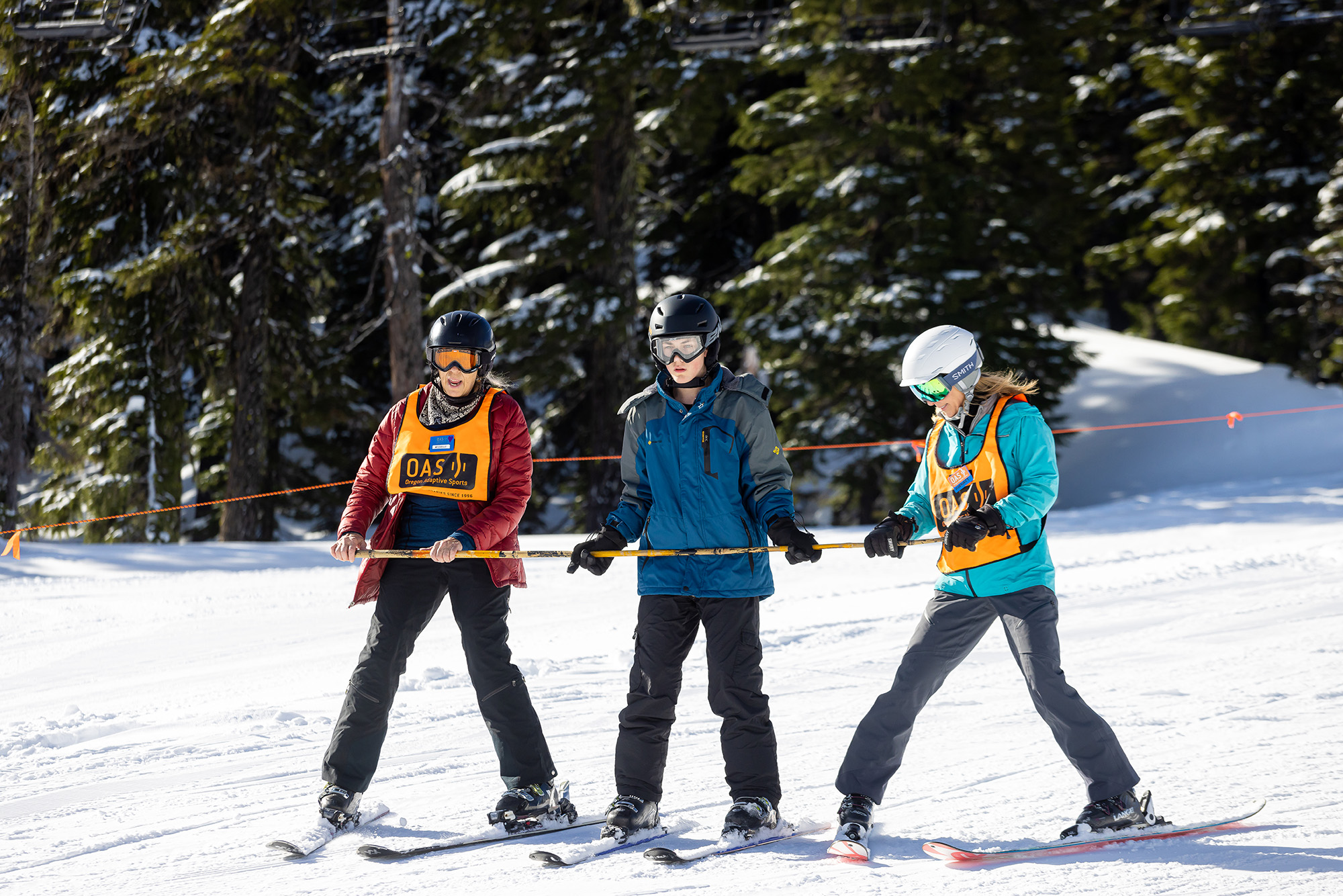 Two OAS volunteers on skis support an athlete between them with a bamboo pole across the three of them.