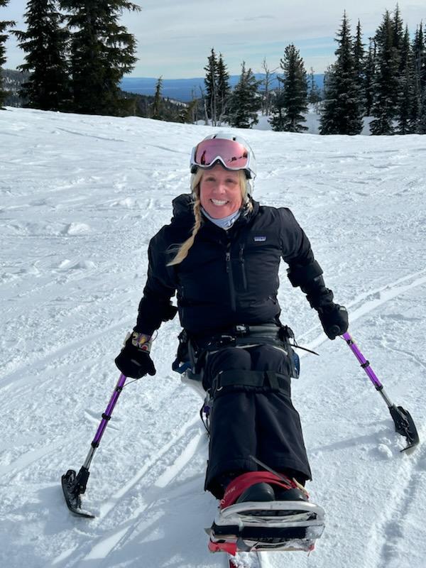 Collen with pink tinted goggles, blonde hair in a side braid in a sit-ski on the slopes.