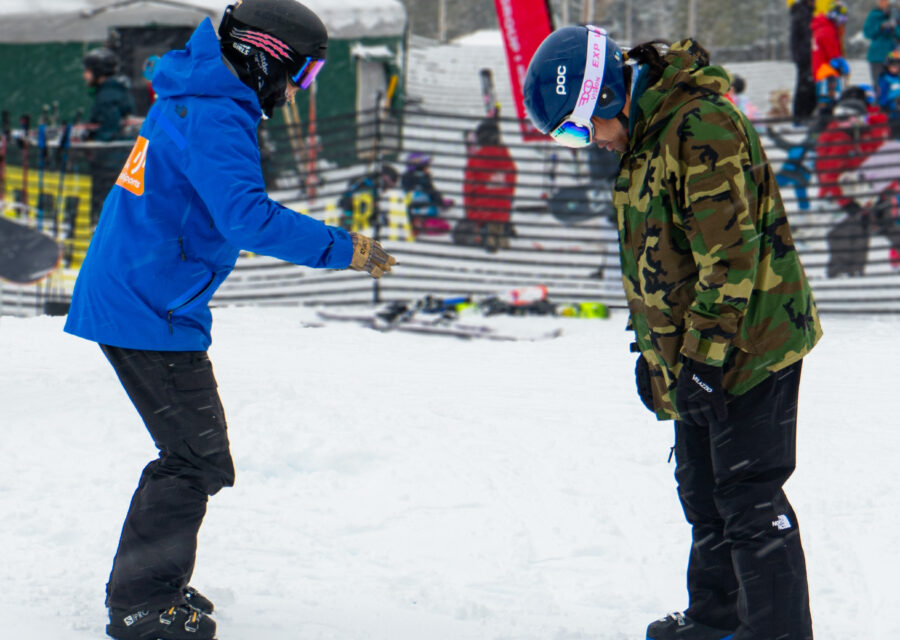 an OAS instructor stands, no skis, facing an athlete, Veteran, who is wearing a camouflage winter jacket