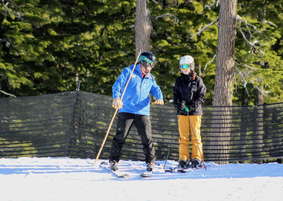 An OAS instructor in a blue jacket holds a long bamboo pole upwards and speaks to a standing skier