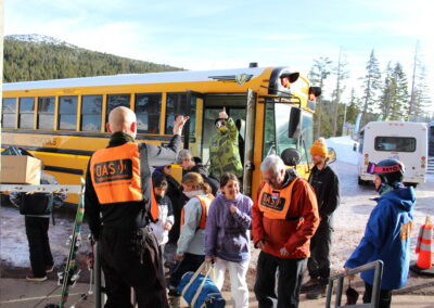 A yellow school bus with students unloading and being greeted by OAS volunteers in orange vests