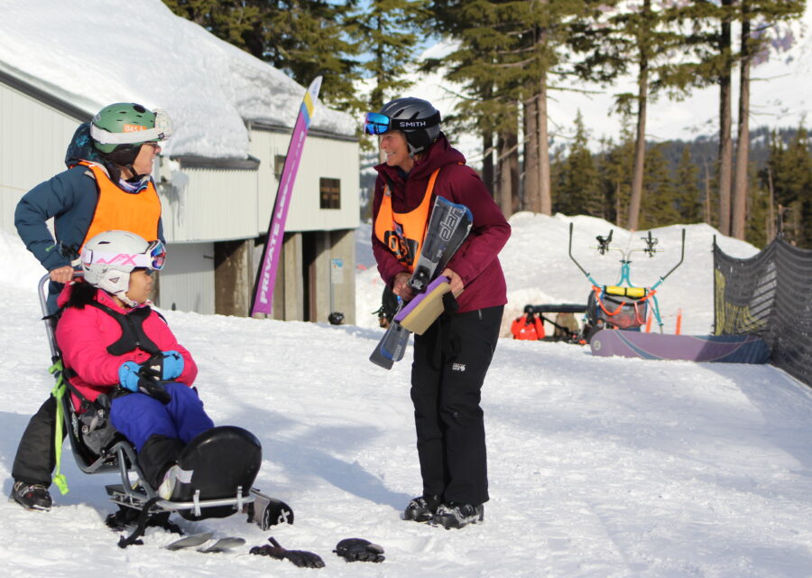 A young athlete in a sit ski is assisted from behind by a volunteer facing another volunteer holding skis