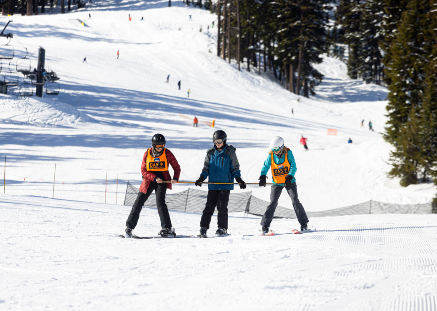 Two OAS volunteers ski beside an athlete using a long bamboo pole to support them