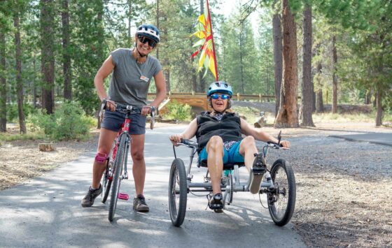 Lorrie wearing a helmet and sunglasses in a recumbent bike on a paved path, beside her OAS stands with their stand up bike