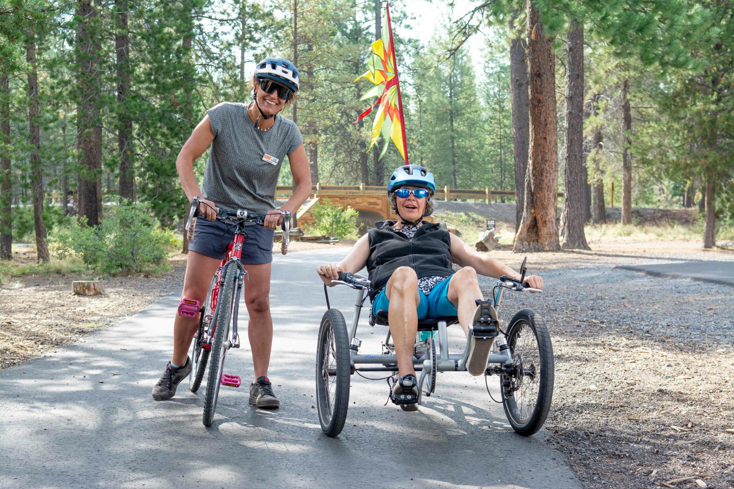 Lorrie wearing a helmet and sunglasses in a recumbent bike on a paved path, beside her OAS stands with their stand up bike