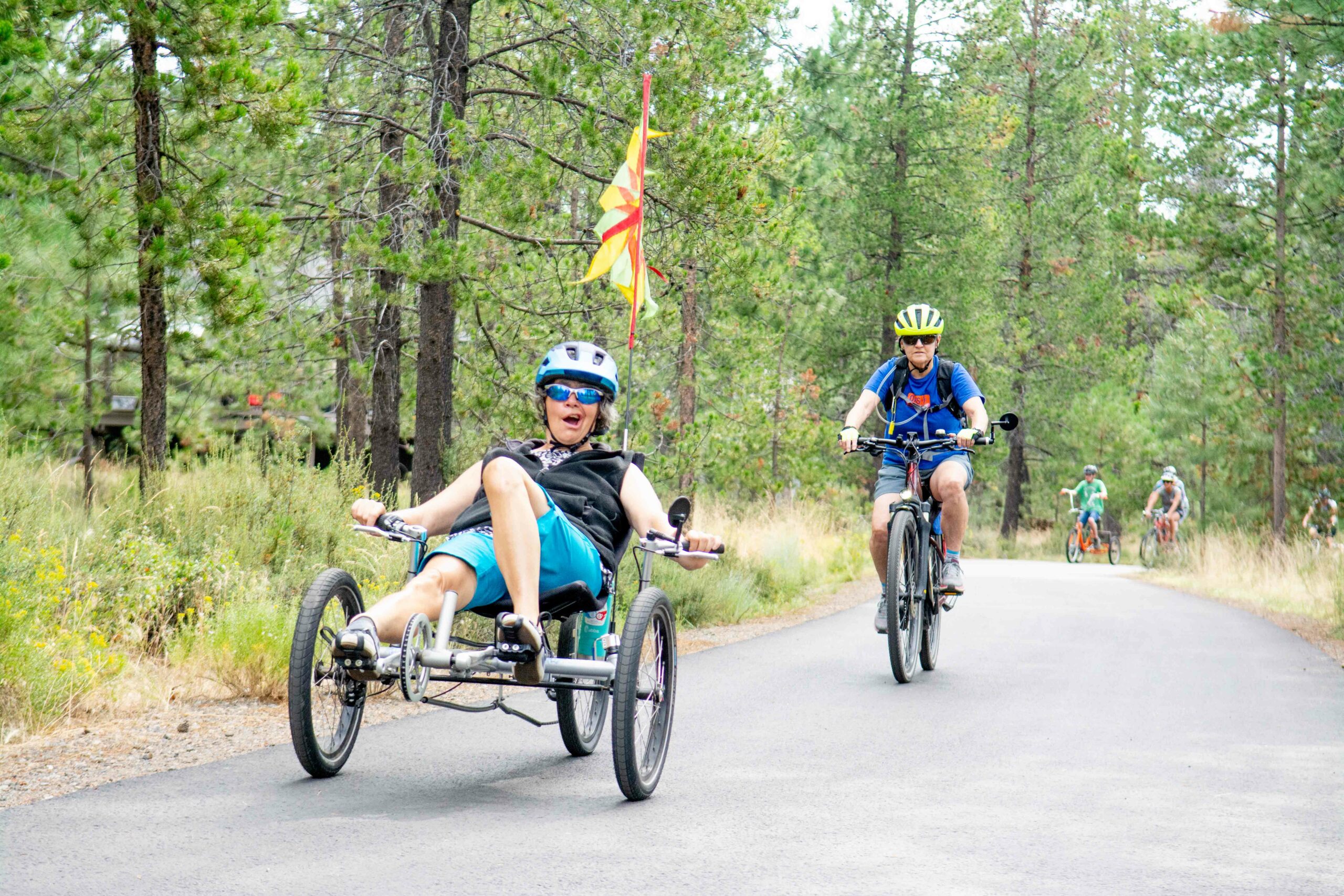 Lorrie pedals in her recumbent bike down a paved path with green trees in the background