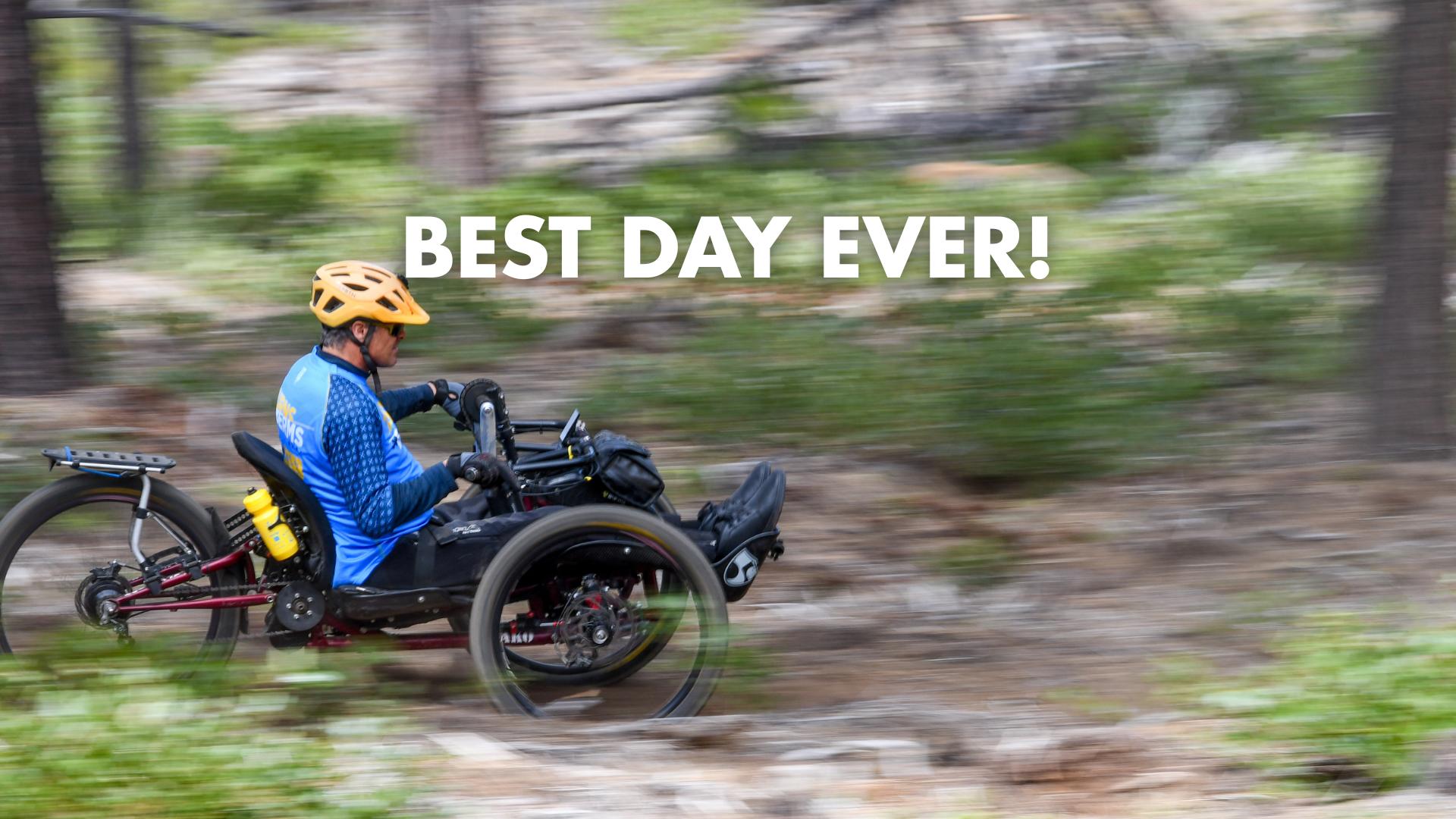 an adaptive mountain biker cruises down a dirt trail, surrounded by greenery. The background is blurry, showing the speed the athlete is moving at.