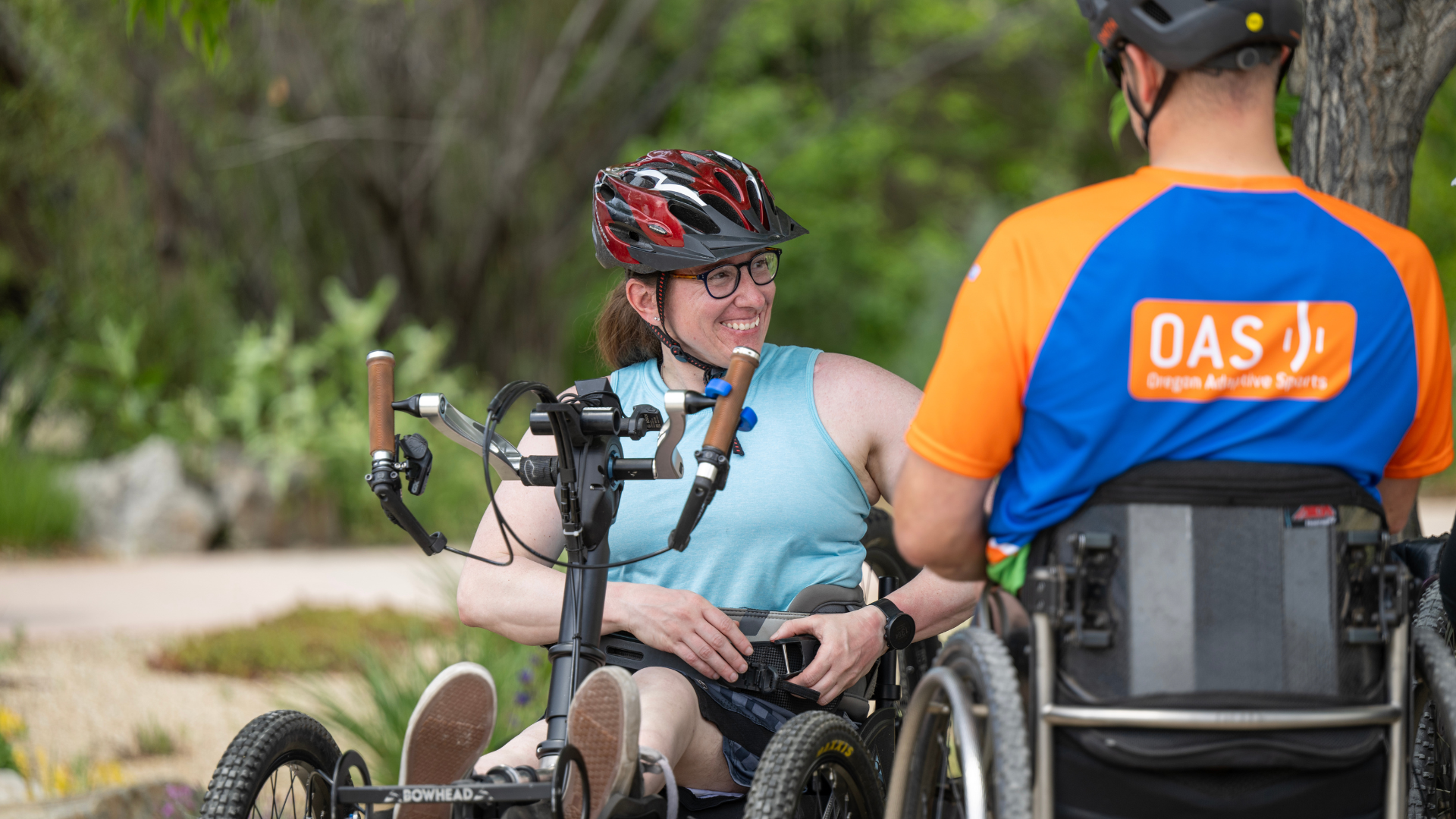 A woman grins at an O-A-S instructor, who is facing away. The woman is wearing a blue tank top and a red helmet, and adjusting equipment at her waist. She is sitting on an adaptive Mountain Bike, and the instructor is using a wheelchair.