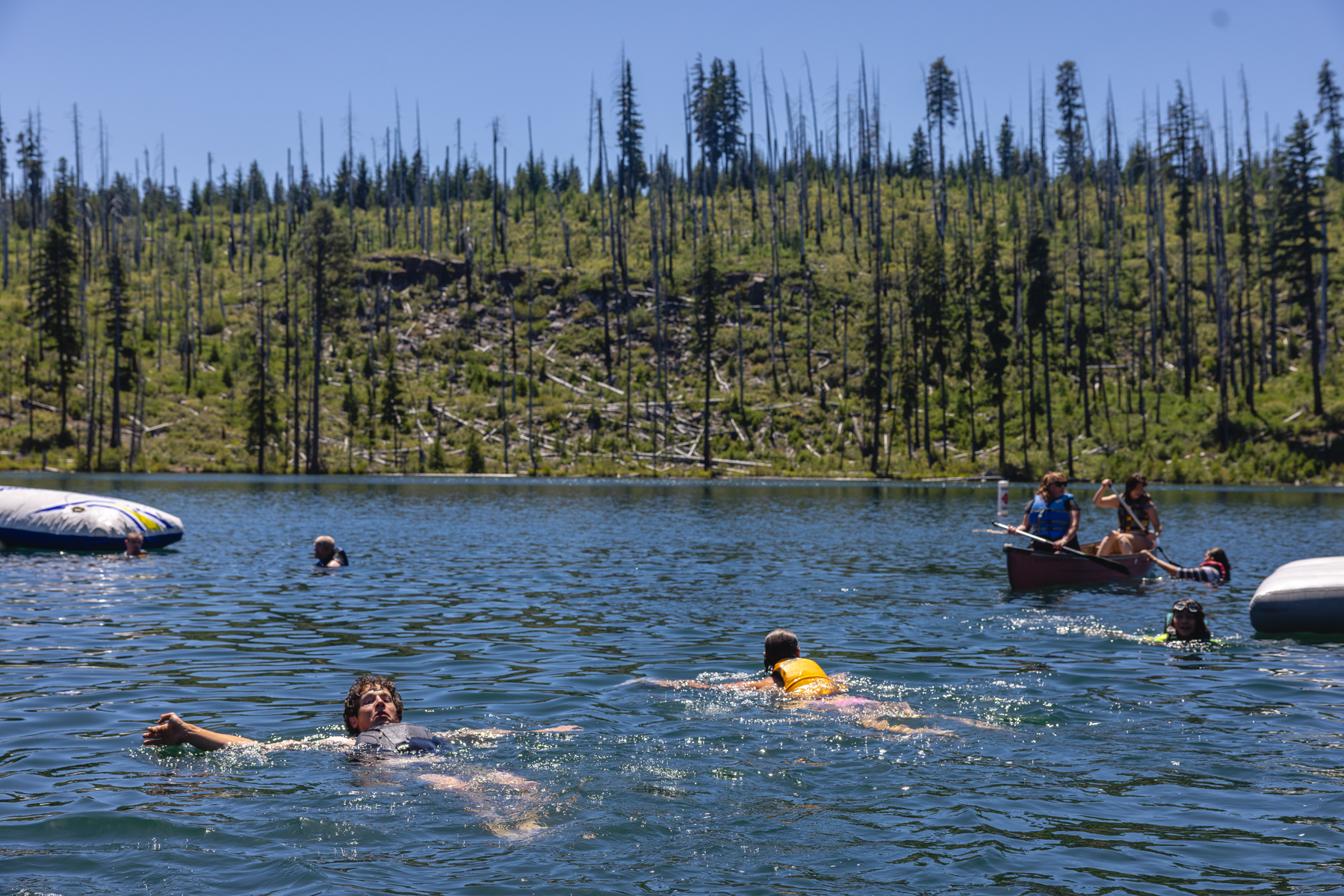 Dave, a man with brown curly hair, floats on his back on Dark Lake in a P-F-D. Around him, others swim, paddle, and float in the water. 