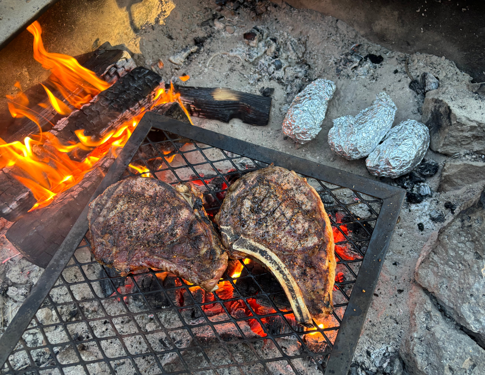 two ribeye steaks cooking on a grill over a campfire