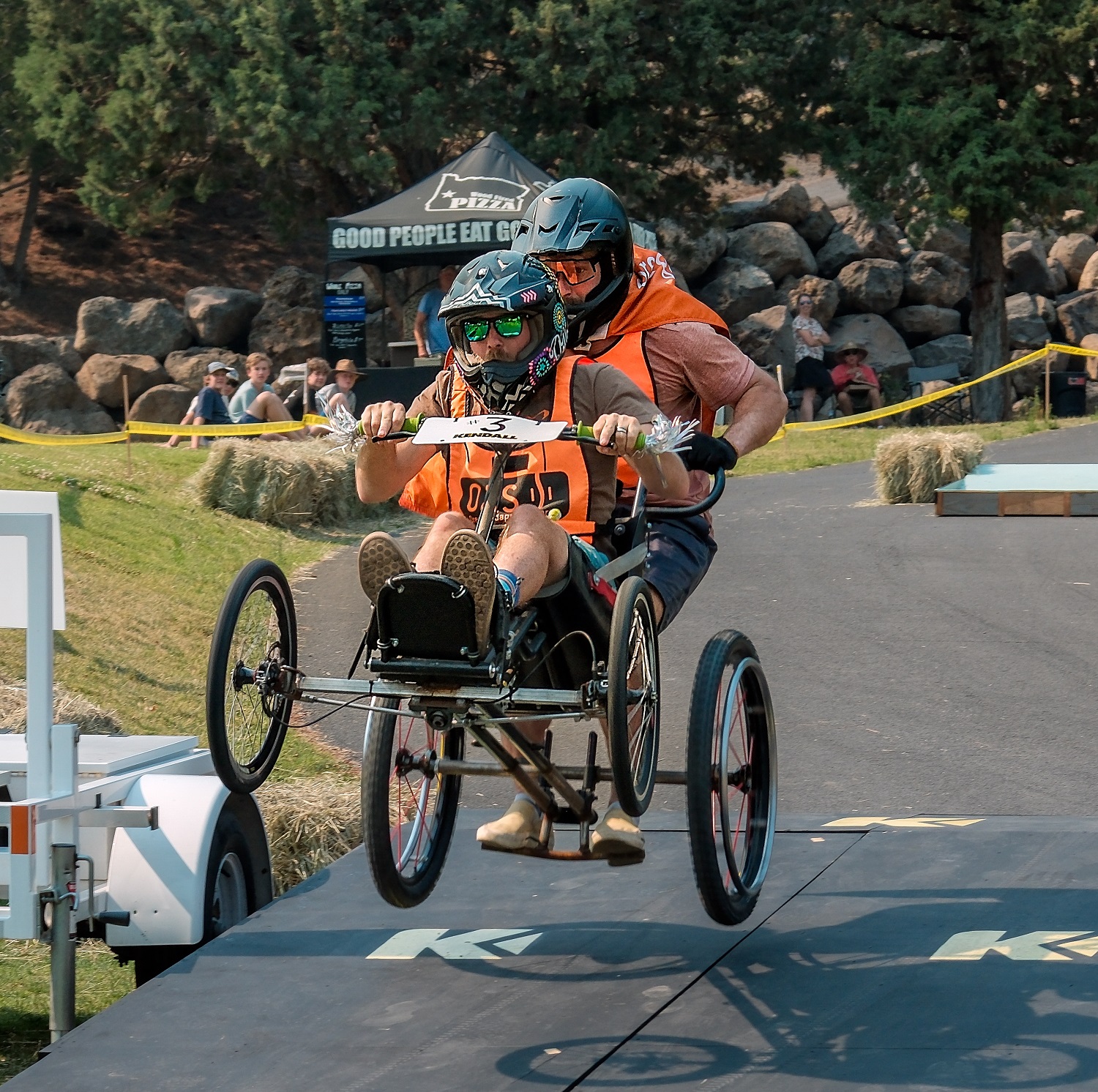 two people wearing bright orange vests with the O-A-S logo on them ride a four-wheeled go-kart over a jump. They are in the air and both wearing full-face helmets. 