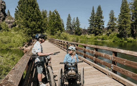 Sophie, a blonde woman riding a Bowhead adaptive mountain bike, stops to chat on a wooden bridge over the river. She chats with Melodie, O-A-S instructor who stands over her bike.