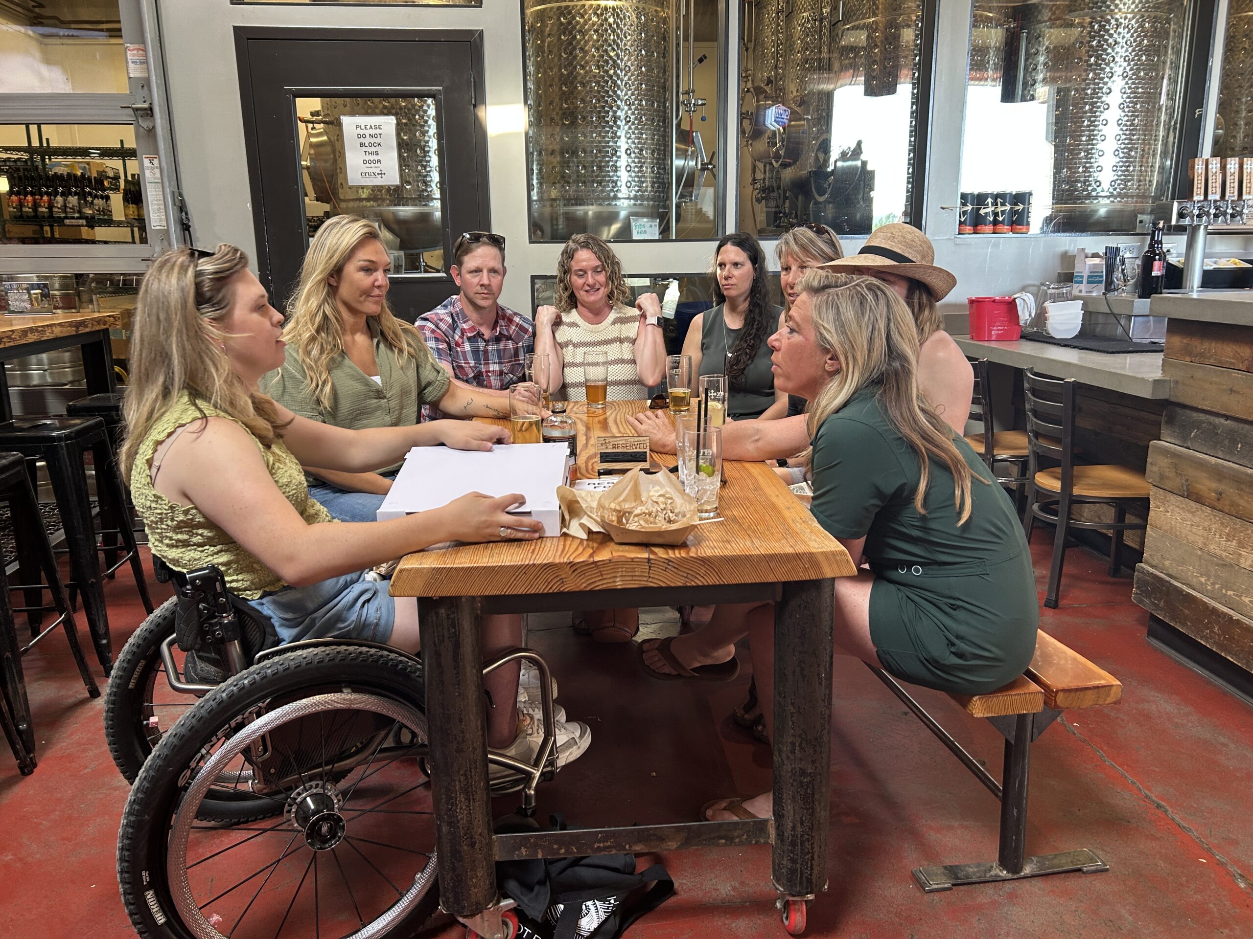 a group of people gather around a wood table at a brewery. 