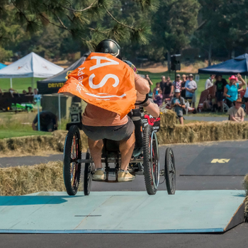 a person standing on the back of a four-wheeled go-kart wearing an orange O-A-S flag 