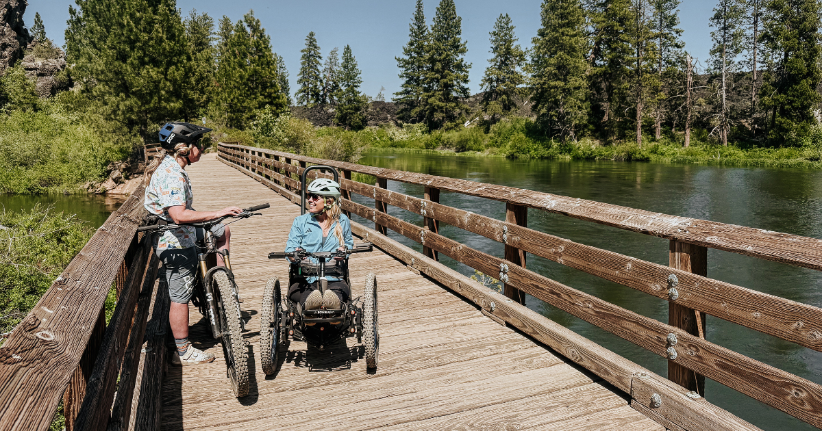 Sophie, a blonde woman riding a Bowhead adaptive mountain bike, stops to chat on a wooden bridge over the river. She chats with Melodie, O-A-S instructor who stands over her bike. 