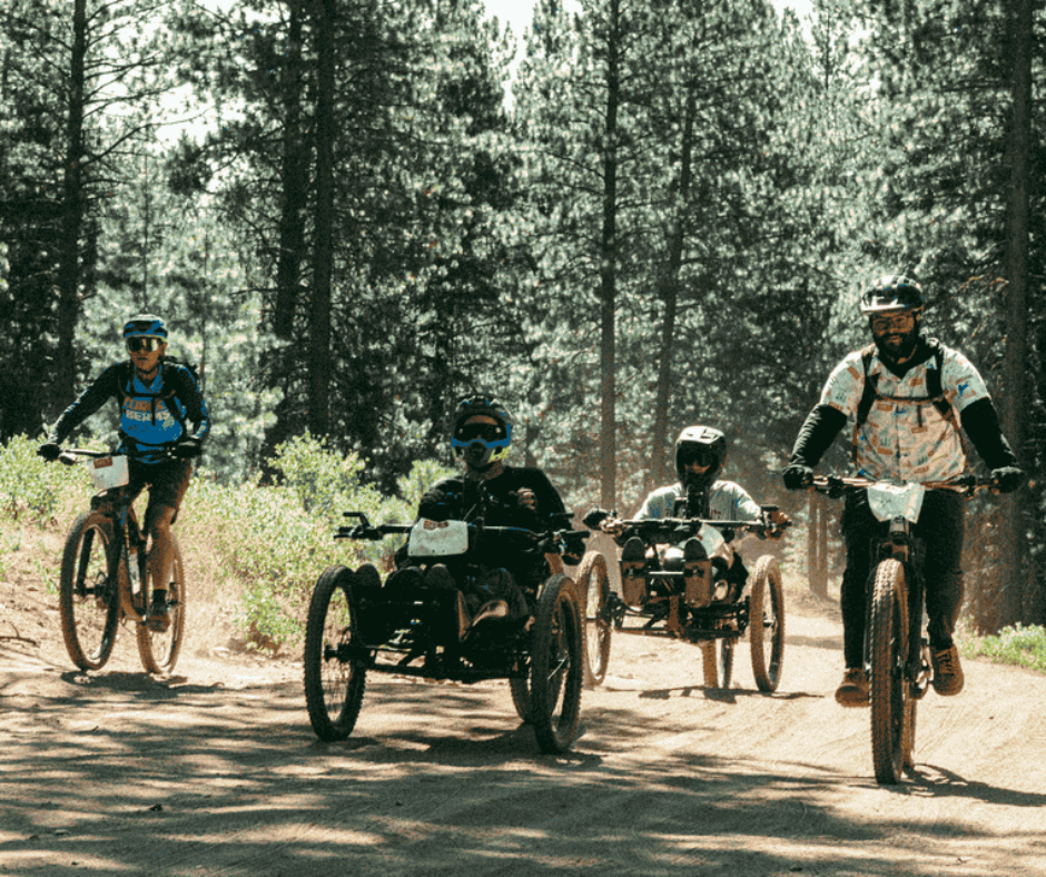 four people on bikes ride down a dirt road. The two on the outsides are on stand bikes and the two in the middle ride adaptive mountain bikes