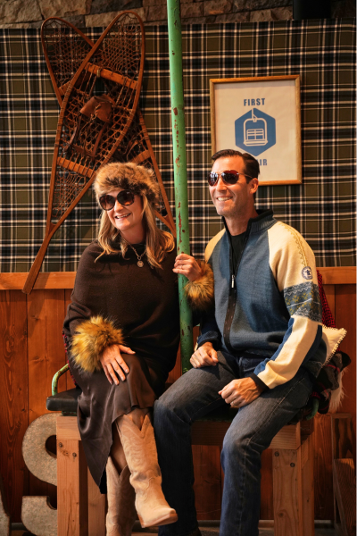 a man wearing aviator glasses and a knit sweater and a woman wearing a fur hat and sunglasses sit on the chairlift photo booth.