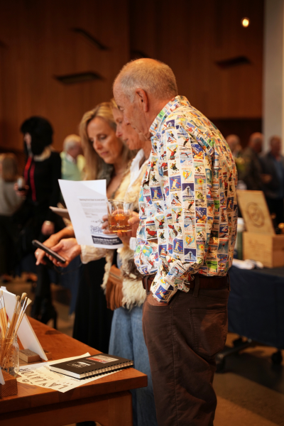 three people looking down at auction display packages. In the front in focus, a man with white hair wears a button down shirt with ski area posters on it.