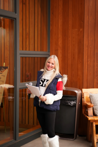 Molly, the First Chair Auctioneer, smiles directly at the camera. She has very light blonde hair, and is wearing a white turtleneck sweater with a fur lined vest and fur lined boots.