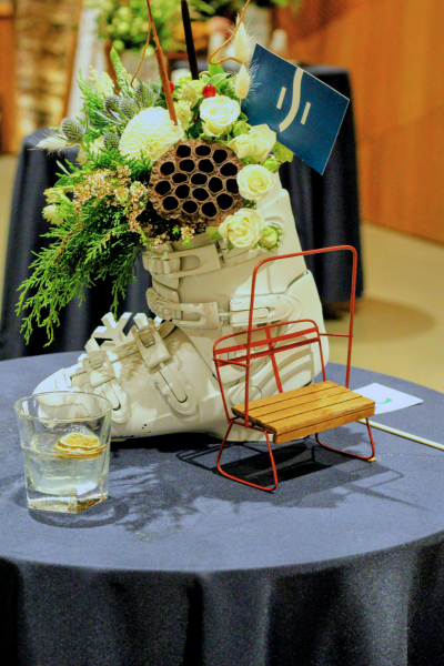 a white ski boot sits on a blue tablecloth. The ski boot is acting as a vase for a flower arrangement, and has a mini red chairlift chair and a cocktail in front of it.