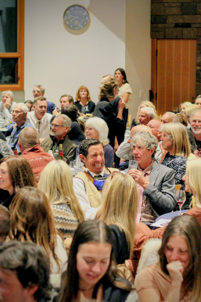 a shot of the dinner tables, filled with people. A man in the center of the room is in focus, with dark hair, a fuzzy white vest, and O-A-S goggles around his neck. He is mid laugh.