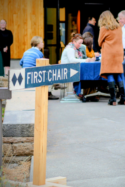 a view of the entrance to the gala: a ski sign with "first chair" and an arrow to the right. Behind the sign, blurry people check in to the event.