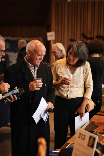 A man wearing a black suit and a woman wearing a white knit sweater with an O-A-S Board member nametag peruse the silent auction packages with drinks in their hand.