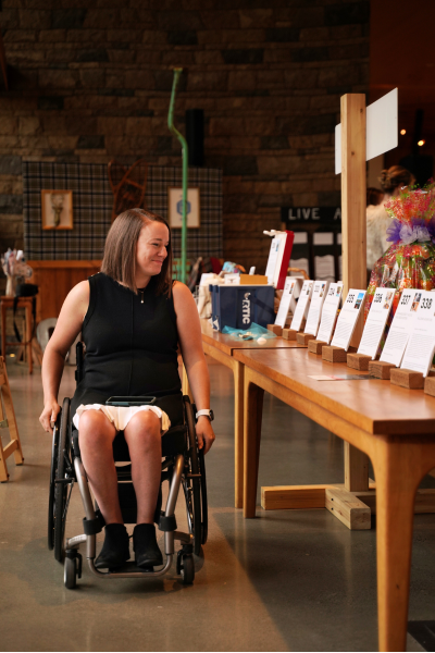 a woman peruses the silent auction before the crowd piles in. She has short brown hair, is wearing a black dress with no sleeves, and navigates the aisle in her wheelchair.