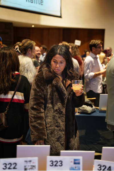 A woman with dark hair and a long brown fur coat peruses the silent auction with her drink.