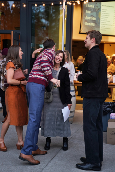 a man in a red knit sweater greets a woman wearing grey and black with a warm hug. Other people around them are smiling.