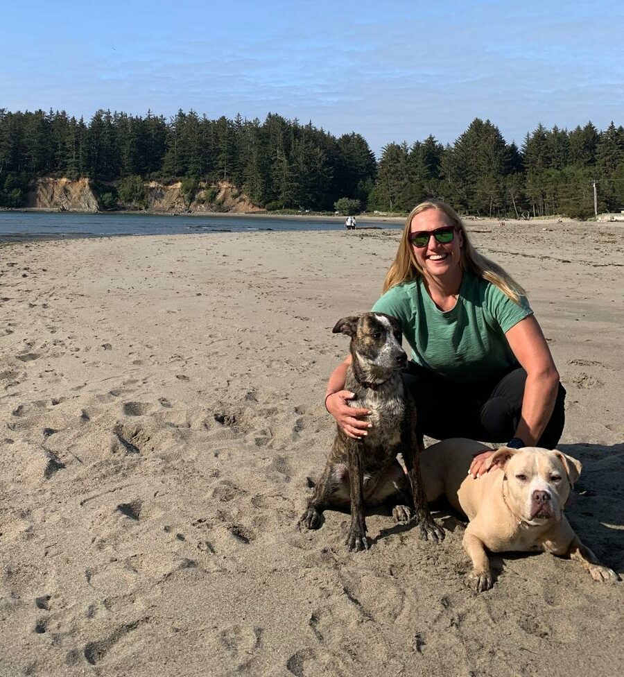 Carly, a white woman with medium lenght blonde hair, kneels on a sandy beach with two dogs. 