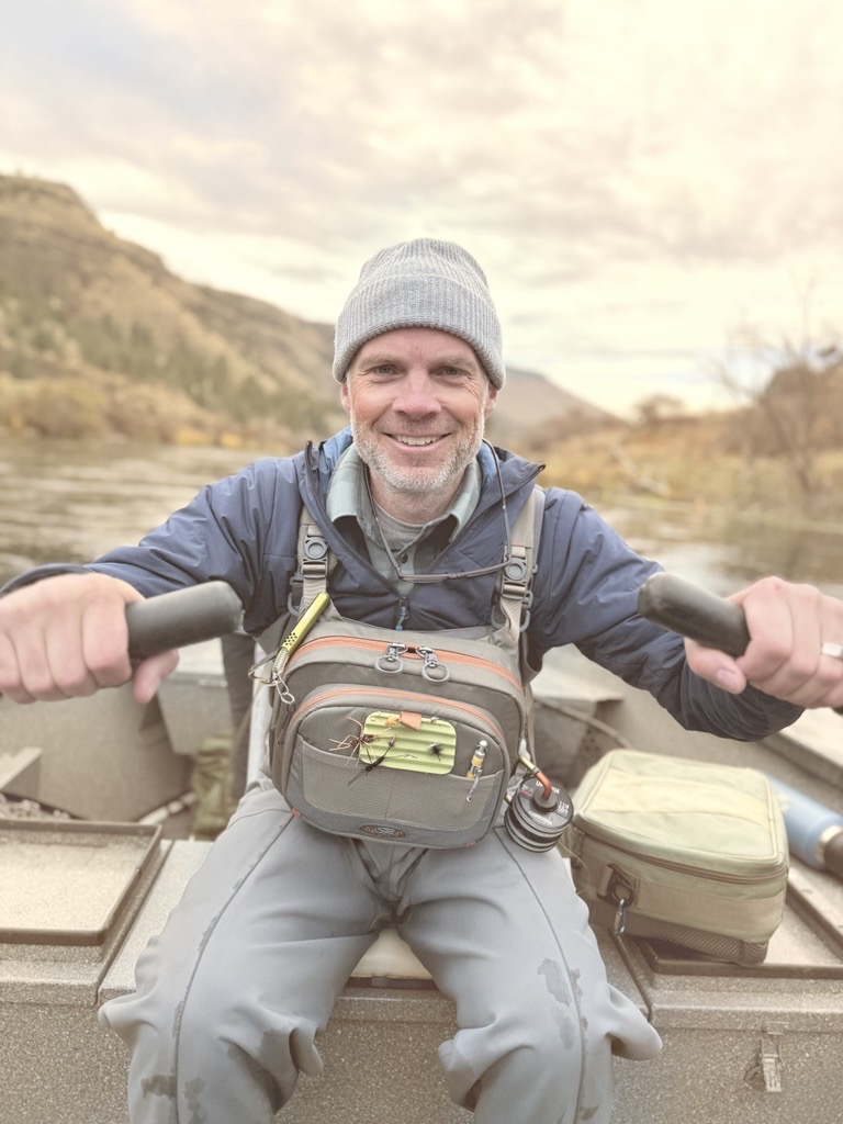 Tim, a white man with silver hair and facial scruff, rows a dory down the river. He wears a fishing P-F-D and grey beanie and smiles at the camera. 