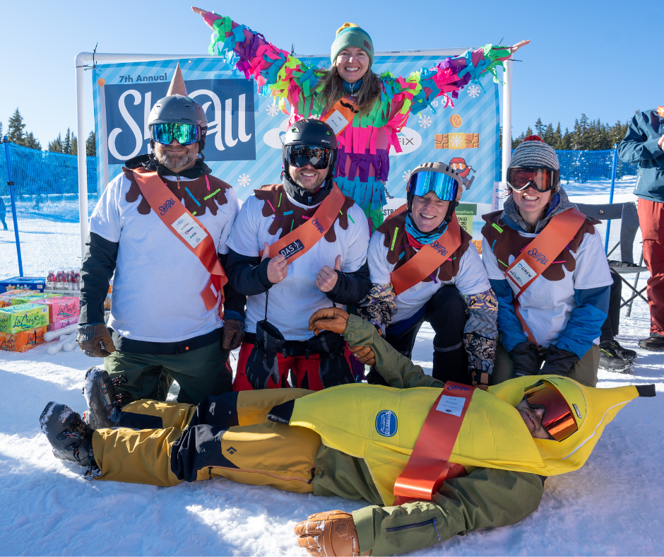 a team dressed up as an ice cream sundae poses for a photo: on the bottom, a person dressed as a banana. Middle row: four people dressed as vanilla ice cream scoops, and on the top, a woman with blonde hair dressed as rainbow sprinkles.