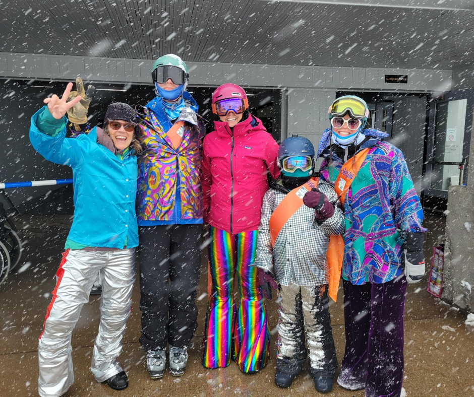 a group of people stand outside sunrise lodge, wearing sparkly costumes while it snows.