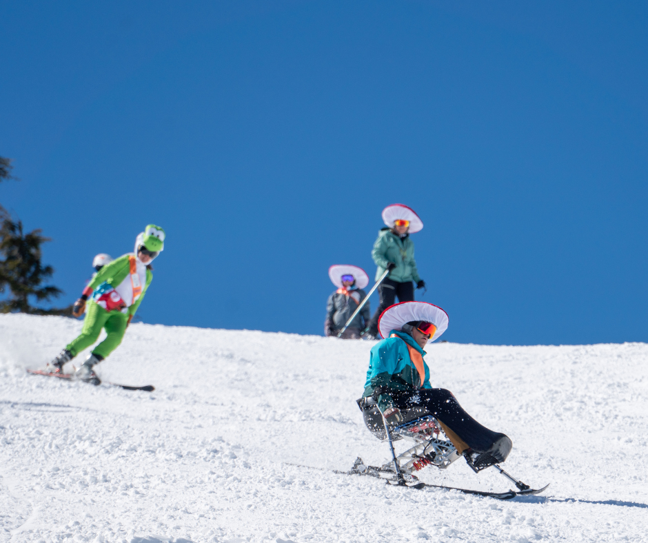 a mono skier in a mushroom hat, a skier in a frog onesie, and a skier in a mushroom hat all make turns on the slope with blue sky behind them.
