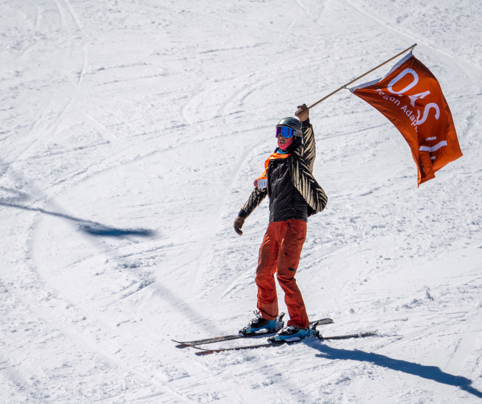 a man wearing a gold sparkly sport coat and red pants carries the orange O-A-S flag while skiing.