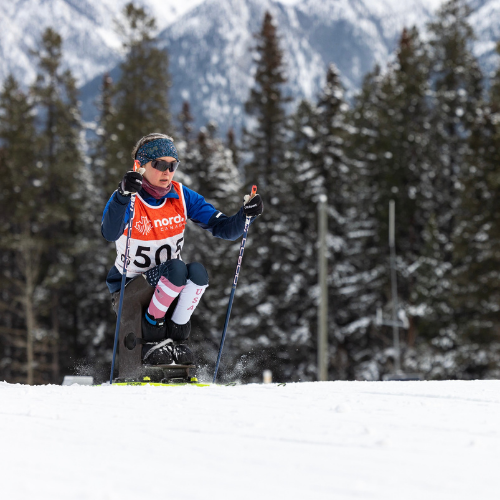 Erin propels herslef forward using nordic poles in a sitski. She wears red, white, and blue and is wearing a red race jersey.
