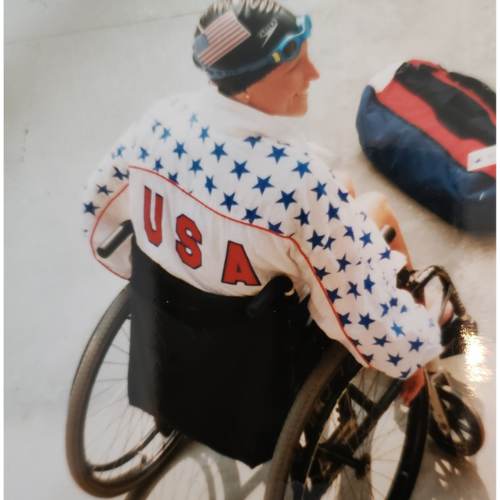 Colleen wears her swim cap and team U-S-A jacket while sitting poolside, in her wheelchair. 