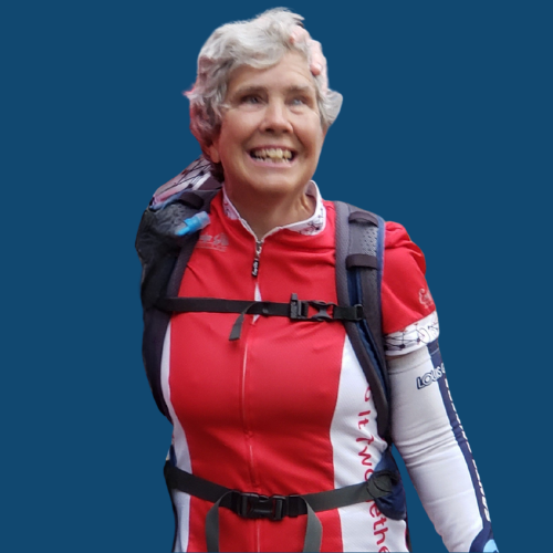 Nancy, a white woman with grey and white hair, stands smiling in a red and white bike jersey. 