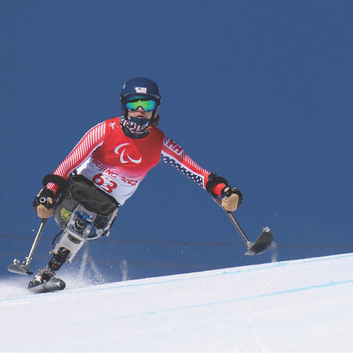 ravi skis his monoski, leaning to the right of screen, with a blue sky in the background. He wears all red, white, and blue and a red race jersey