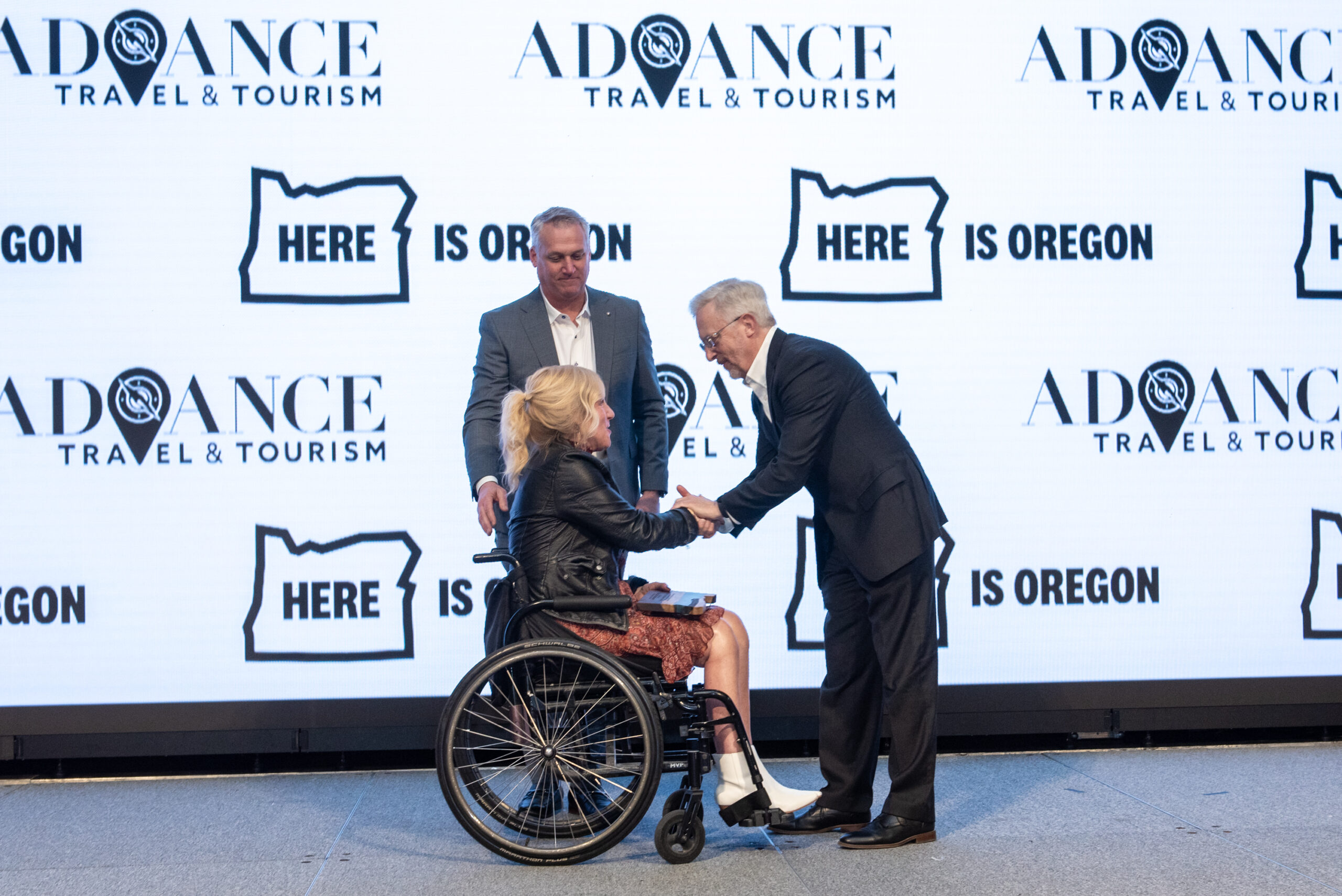 A blonde woman in a leather jacket and sitting in a wheelchair accepts an award on stage from a man with white hair wearing a tuxedo. Another man in a grey suit stands behind them.