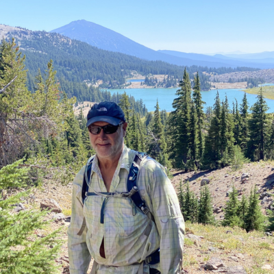 Mountain landscape with lake and forest; a man wearing a hat, sunglasses, and a backpack stands on a rocky hillside facing the camera.