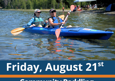 Friday, August 21st. Community Paddling. Paddle and Play. Two people paddle a blue tandem kayak across a lake, each holding a double-bladed paddle, with a forested shoreline