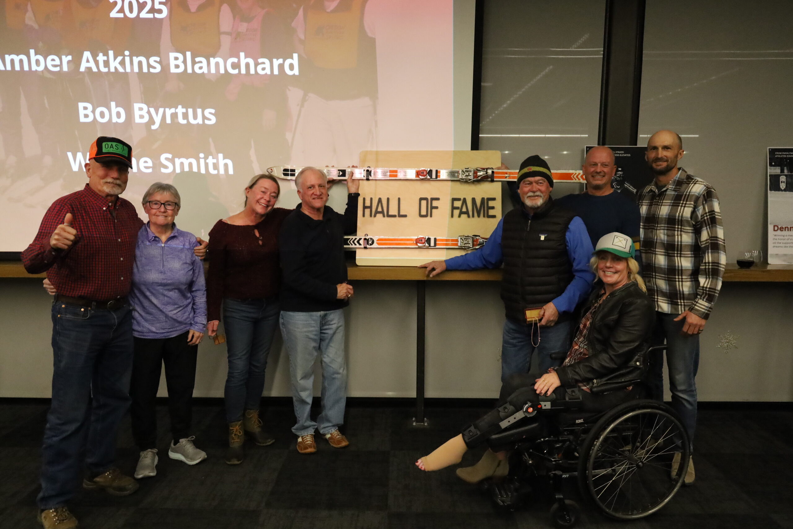 eight people stand together smiling while one person sits in a wheelchair at the front right, with several people holding a sign that reads “HALL OF FAME”; visible text on the screen behind includes “2025” and a list of names.