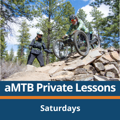 aMTB Private Lessons. Saturdays. A person rides a three-wheeled adaptive mountain bike over a rock feature on a forest trail while another person stands nearby spotting, with trees and rocks in the background.