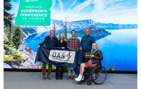 Event photo with a scenic lake and mountain backdrop; five people pose together, four standing and one seated in a wheelchair holding a wooden award, while two people hold an OAS banner across the front; a green graphic in the upper left reads “Oregon Governor’s Conference on Tourism.”