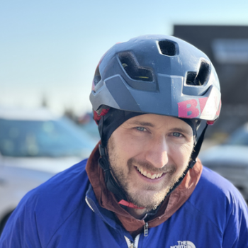 Outdoor close-up portrait; Jesse smiles at the camera wearing a black helmet with “B4” on the side, a blue jacket, and a brown hooded layer underneath, with parked cars blurred in the background.