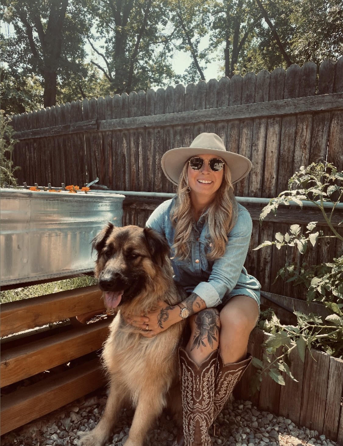 Backyard garden scene; Taylor sits on a wooden bench smiling while holding a large dog at her side, wearing a wide-brim hat, sunglasses, a denim shirt dress, and cowboy boots; a metal planter box and plants sit beside her with a wooden fence behind.