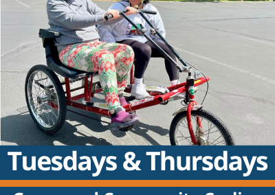 Tuesdays and Thursdays. Group and Community Cycling. Pine Nursery Park. Two people wearing helmets ride a red side-by-side adaptive cycle on a paved parking lot, both smiling and holding the handlebars, with parked cars and trees in the background.