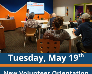 Classroom scene; a group of adults sit at tables facing a presenter standing near a screen at the front of the room; the presenter gestures toward the screen while speaking; text at the bottom reads “Tuesday, May 19th” and “New Volunteer Orientation.”